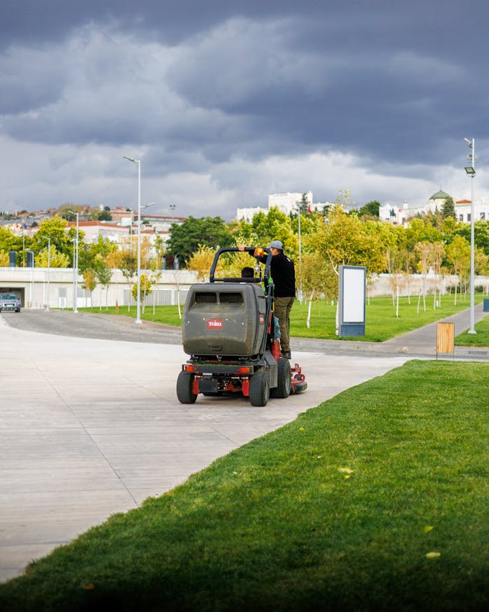 A park employee driving a lawnmower on a cloudy day in an urban park setting.